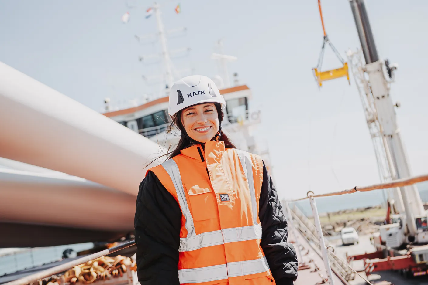 Cátia Alves on a quay in high-visibility vest and helmet, beside a wind turbine blade, with a vessel and crane behind her